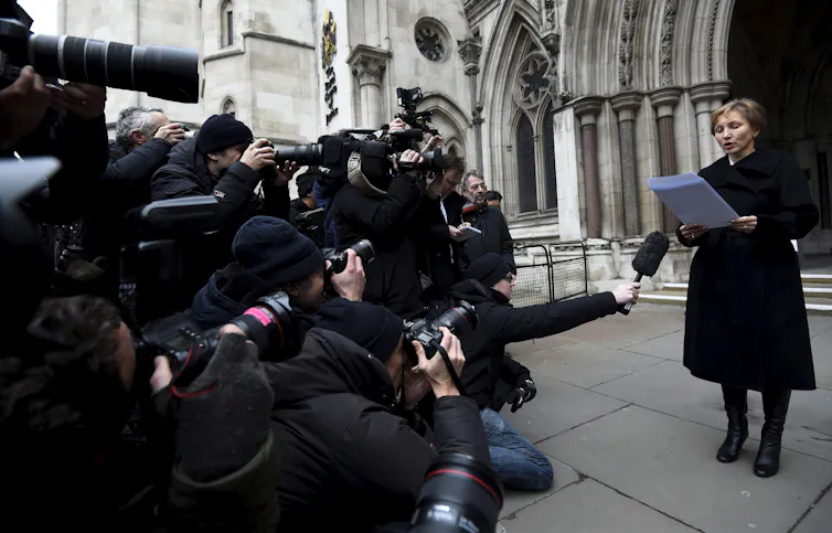 A woman wearing all black reads a statement in front of a large crowd of photographers and reporters