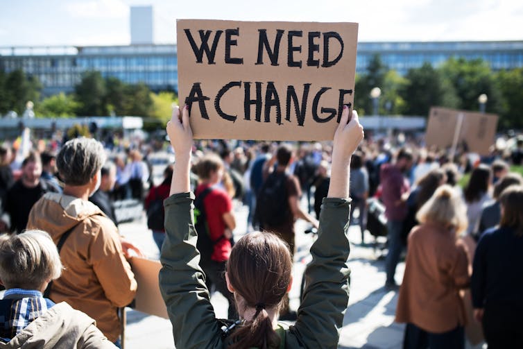 Young person photographed from behind holding a sign saying WE NEED A CHANGE