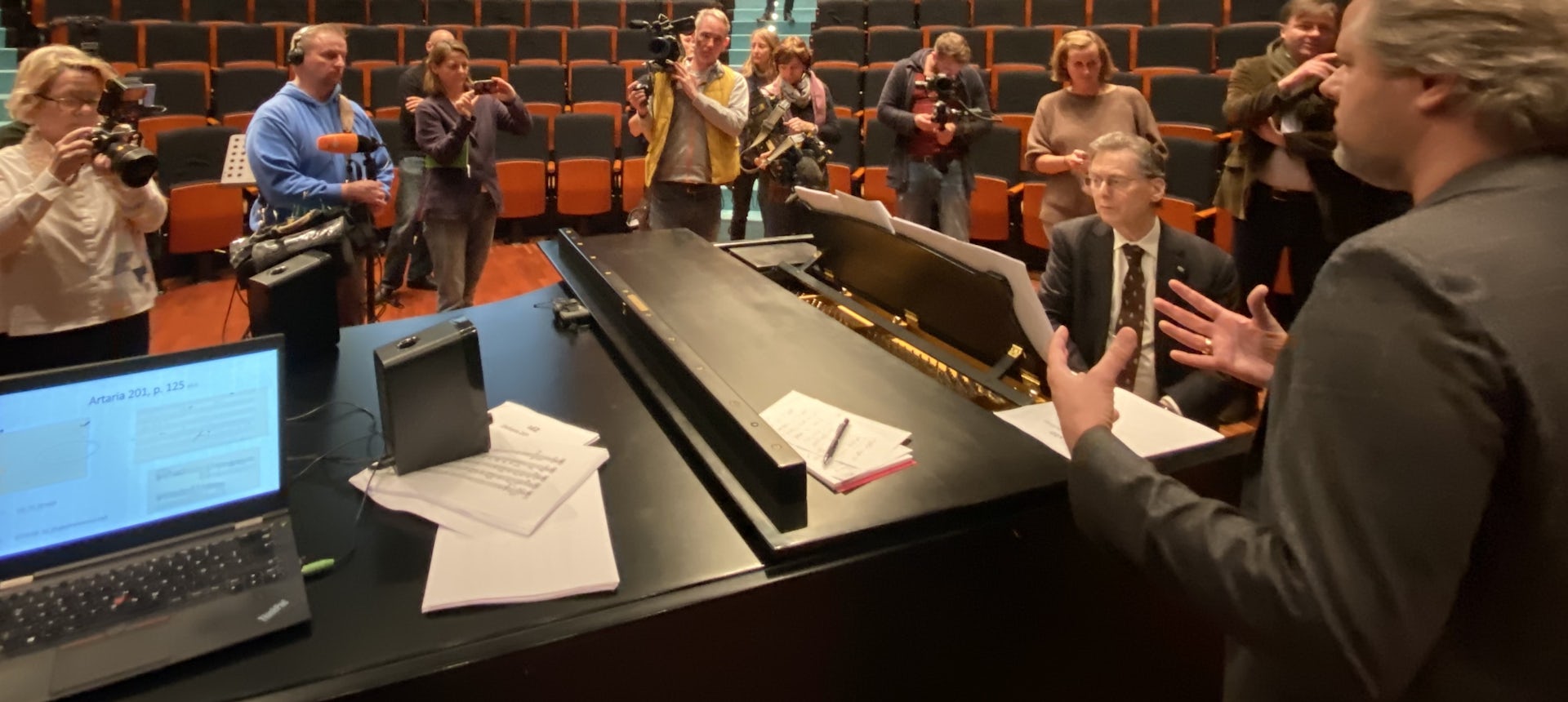 Group of people stand around a piano player in a concert hall