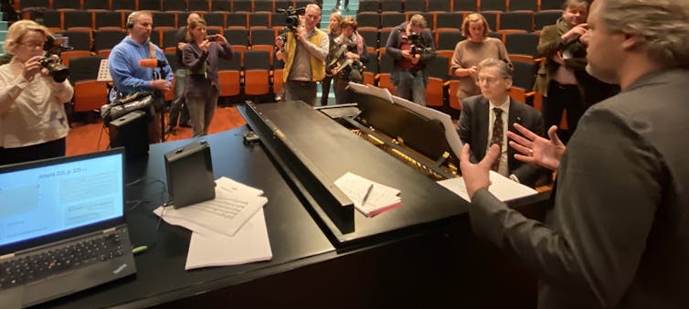 Group of people stand around a piano player in a concert hall