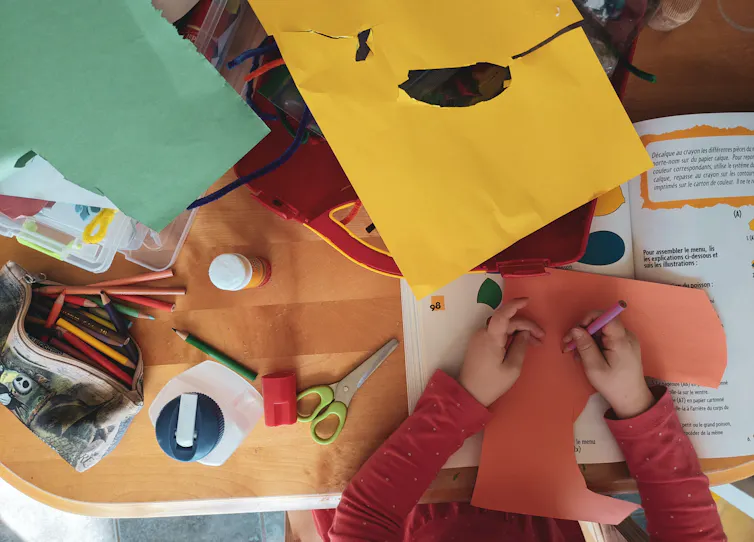 A child holds a pencil crayon to a cut piece of orange construction paper, the photo is from the top-down and the desk is covered with other craft materials