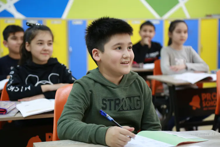 Children sit in a classroom, smiling up at the front of the room