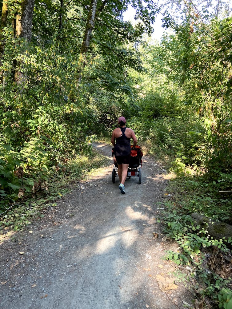 A woman seen from behind, jogging on a wooded path while pushing a double stroller