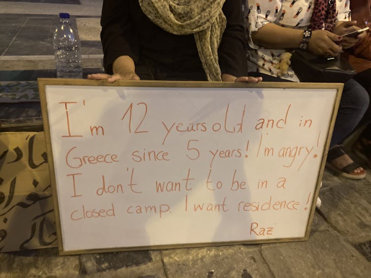 A whiteboard held by a young girl reads 'I'm 12 years old and in Greece since 5 years! I'm angry! I don't want to be in a closed camp, I want residence!'