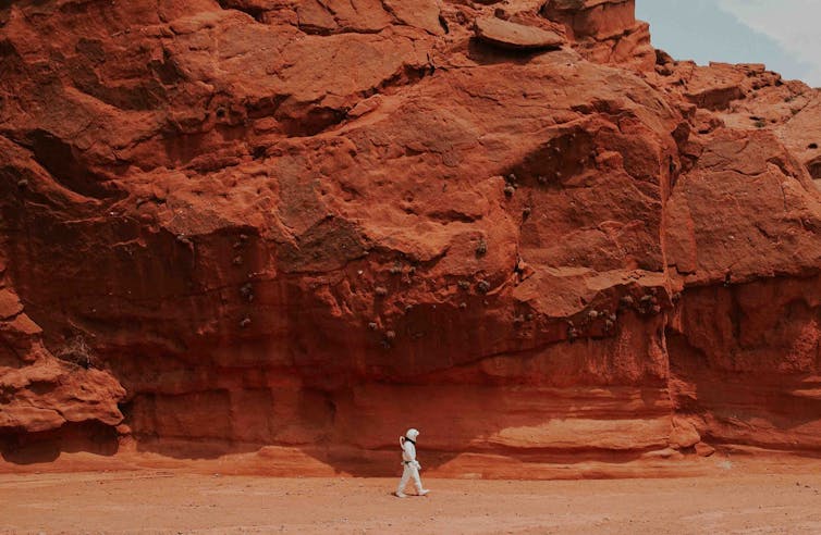 A man in a spacesuit walks across a Martian landscape.