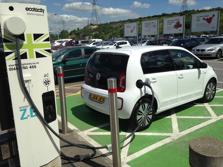A small, white electric car in a green parking bay connected to a charging point.