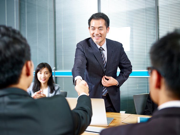 Scene from a Chinese boardroom. Suited executive shakes hands with another businessman as a woman looks on.