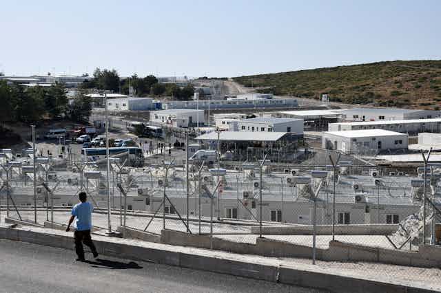 A man is seen walking by a migration centre in Greece, cameras line the fence facing both in and out