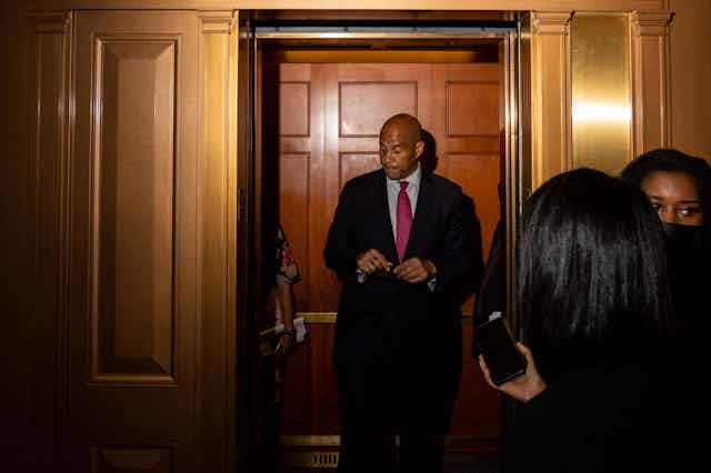 A downbeat looking Senator Cory Booker stands in front of a closed door while journalists wait nearby.