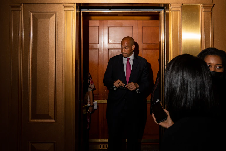 A downbeat looking Senator Cory Booker stands in front of a closed door while journalists wait nearby.