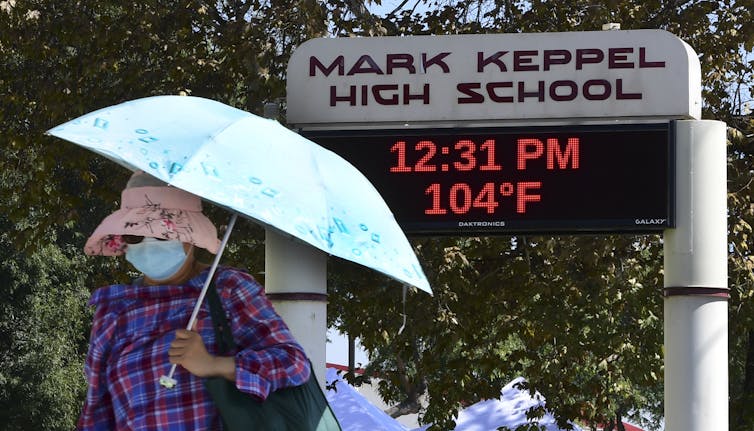 Woman wearing hat and holding umbrella walks by school sign showing temperature is 104 degrees