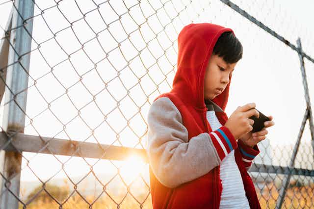 A boy wearing a hooded sweatshirt leans back against a chain-link fence as he holds a smart phone horizontally in both hands and stares at its screen