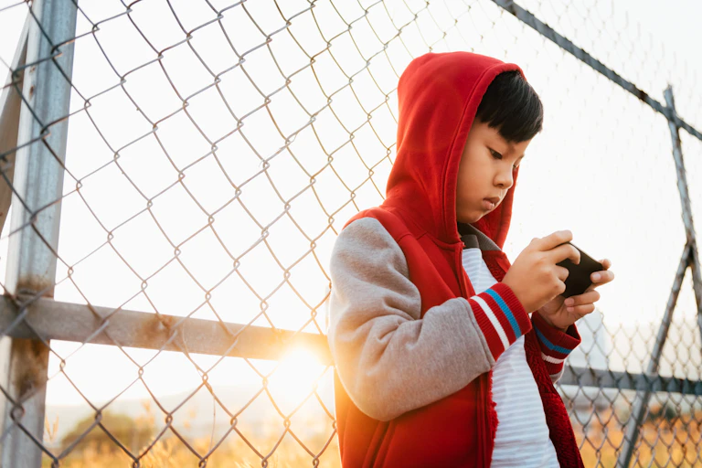 A boy wearing a hooded sweatshirt leans back against a chain-link fence as he holds a smart phone horizontally in both hands and stares at its screen