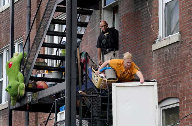 A woman carries a laundry basket full of stuff and a wall picture down a fire escape as a member of the St. Louis sheriff's department watches her eviction.
