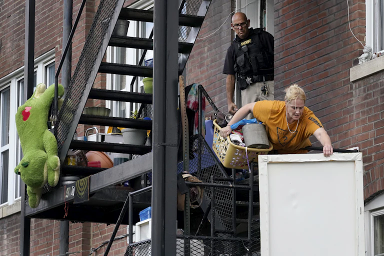 A woman carries a laundry basket full of stuff and a wall picture down a fire escape as a member of the St. Louis sheriff's department watches her eviction.