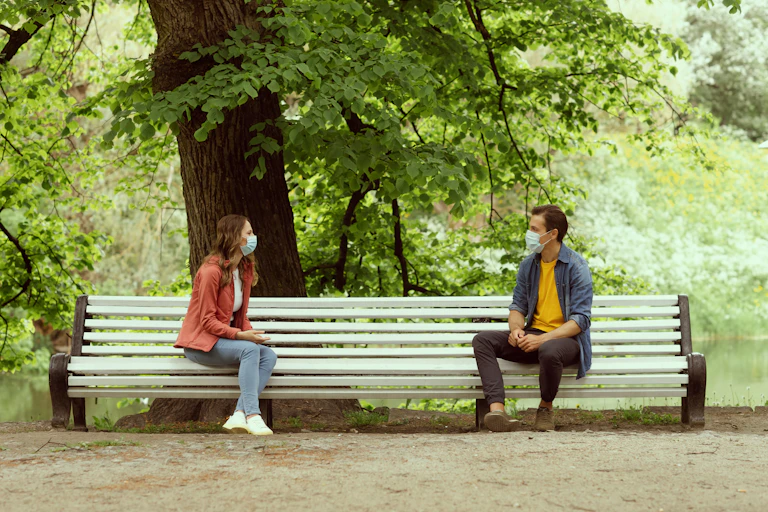 Masked peoepl talking to each other on a park bench.