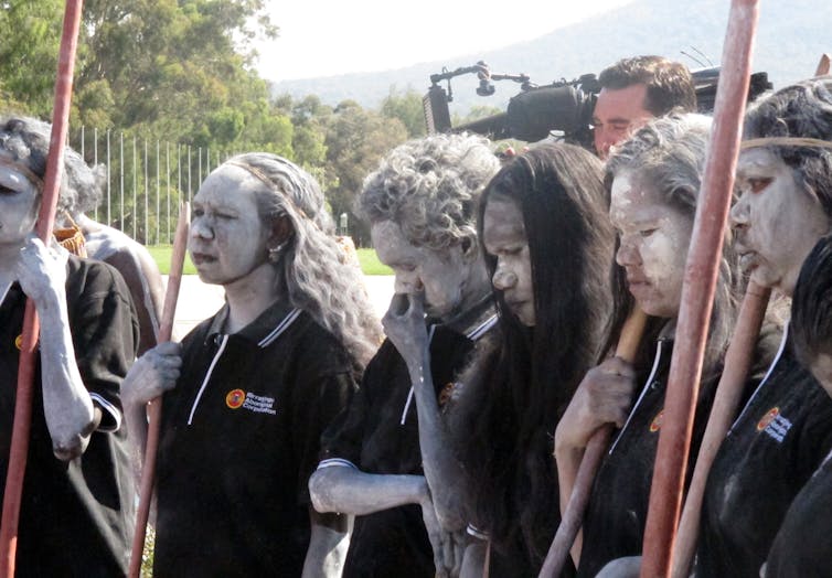 Aboriginal people painted in white ochre, standing outside parliament house.