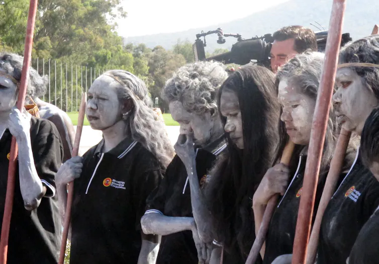 Aboriginal people painted in white ochre, standing outside parliament house.