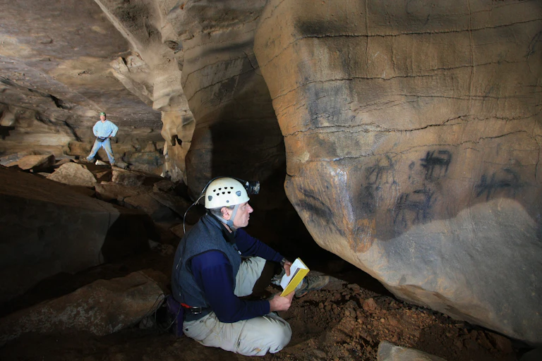 man seated on cave floor looking at black images on stone wall