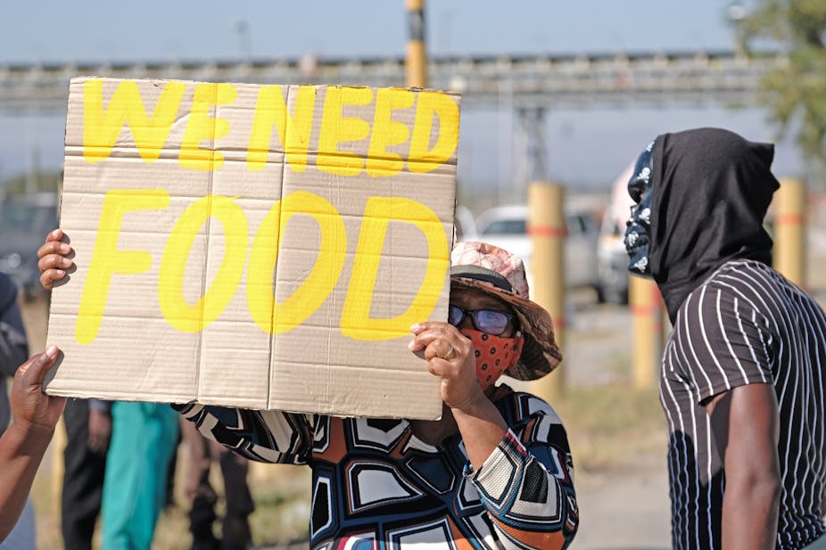 A woman wearing a brightly patterned shirt, a floppy hat, a mask and sunglasses holds up a piece of cardboard that has the words "We need food" written on it in yellow.