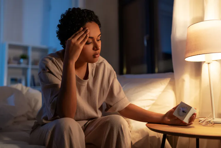 A woman sits on the edge of her bed, looking at her bedside clock.