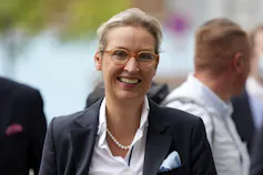 A woman smiling in a blazer, glasses and pearls.