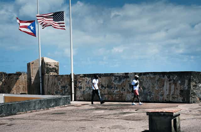 People walk along a concrete sidewalk next to the flags of the U.S. and Puerto Rico