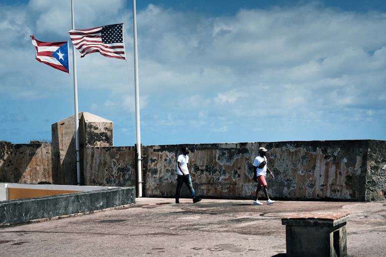 People walk along a concrete sidewalk next to the flags of the U.S. and Puerto Rico