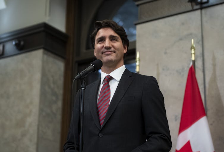 Justin Trudeau smiles from behind a microphone with a Canadian flag behind him.