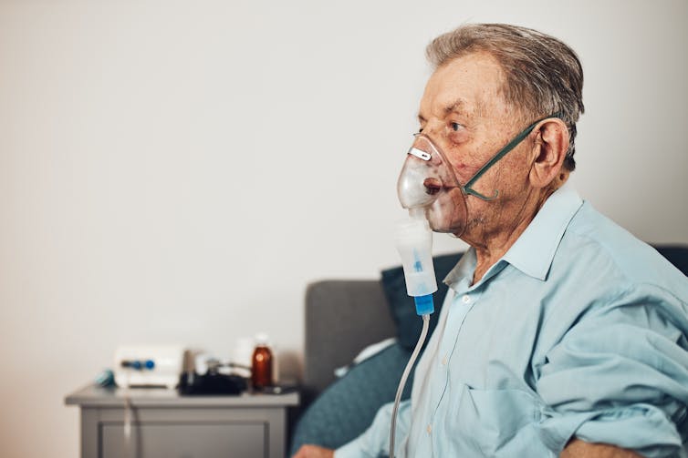 Older man sits on the edge of his bed, breathing through a nebuliser mask.