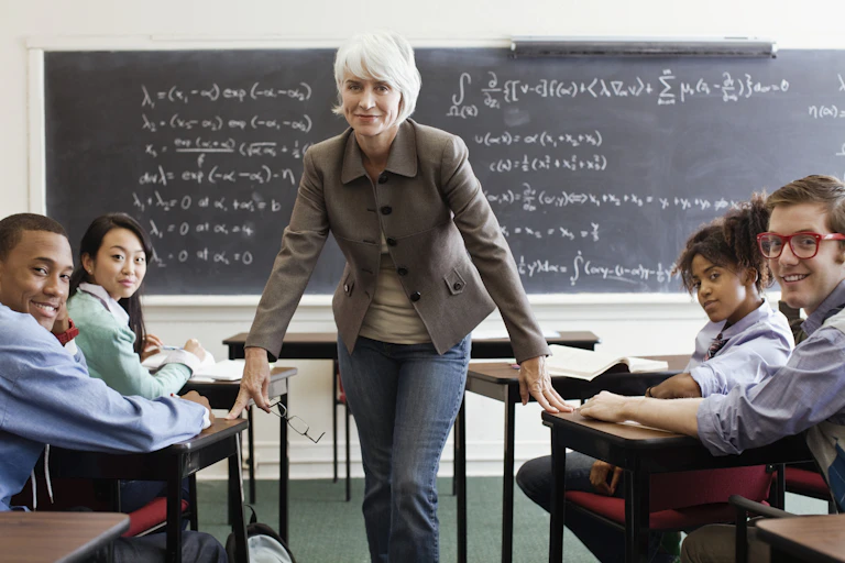 A female teacher in a lecturer room. She's stating between two rows of desks with students seated.