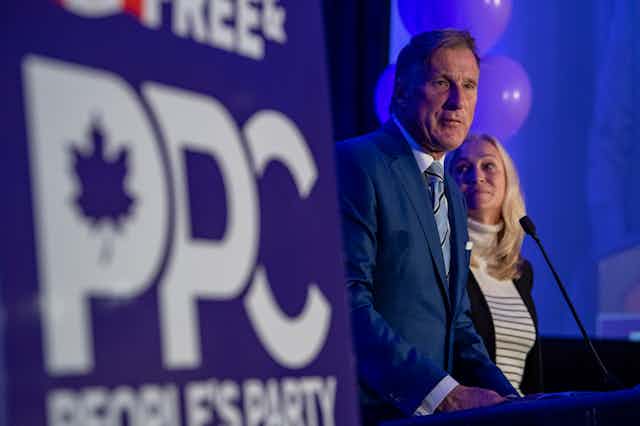 Maxime Bernier in a blue suit speaks to supporters from a podium with a PPC sign next to him. 