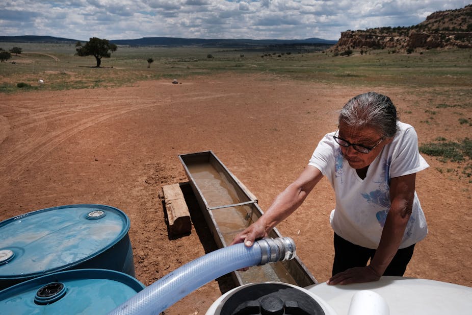 An older woman connects a hose to water tanks with a dry landscape behind her.