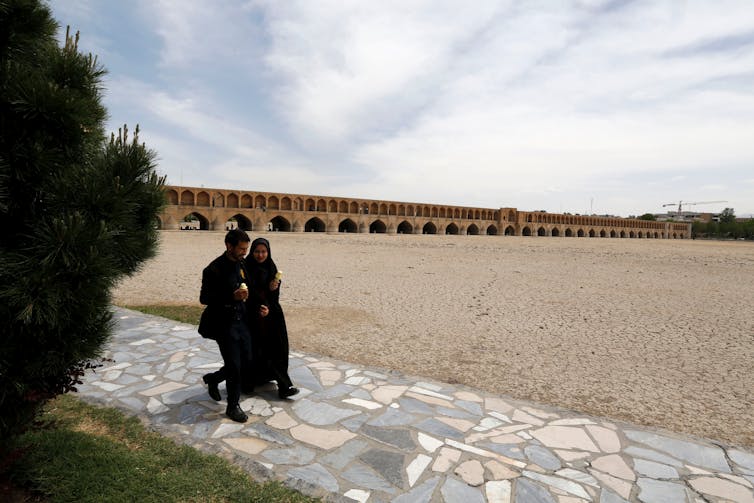 A couple walk on what was once a riverside path. The river bed is dry and cracked.