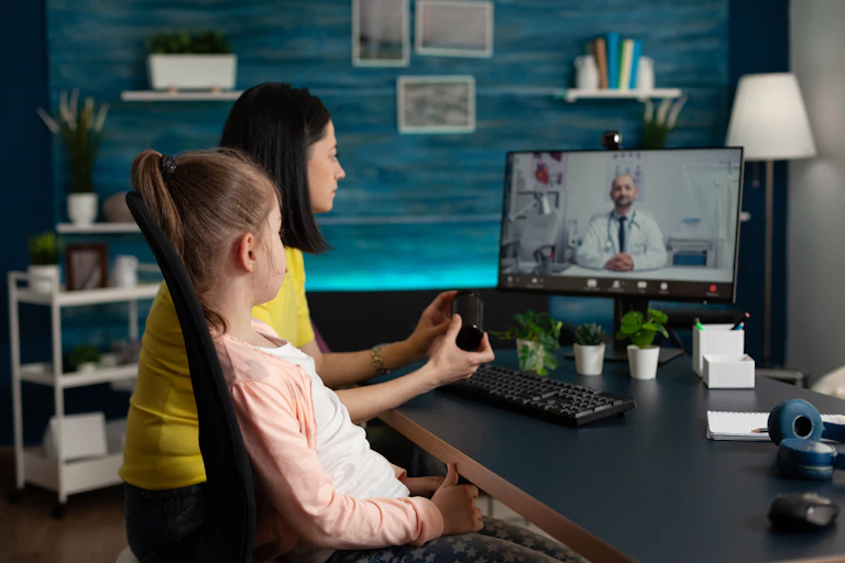 A mother and child sit in front of a computer screen showing a doctor in a white coat