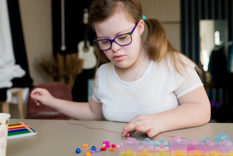 A disabled teen in glasses sits at a desk, beading a necklace.