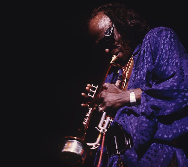 Man in sunglasses dressed in purple playing trumpet.