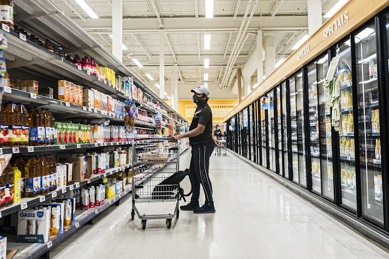 A person with a shopping cart in a grocery store aisle with another shopper in the background