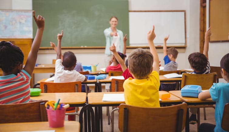 Children sit in a classroom, raising their hands.