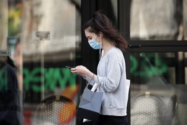 A young woman wearing a mask walks past a building looking at her phone.