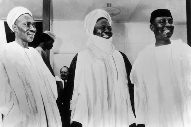 Three men standing and holding hands. The first two from the left are wearing head turbans.