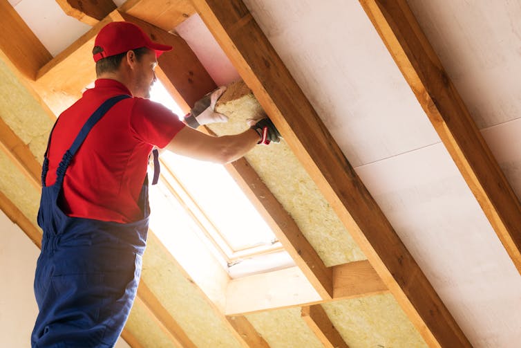 A worker in blue overalls fits wool insulation between attic roof beams.