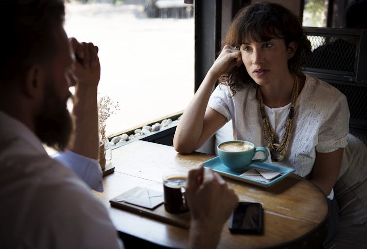 A woman listens to a man speaking in a coffee shop with a latte in front of her.