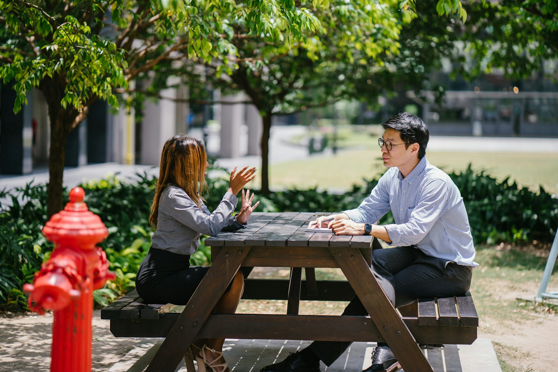 A man and woman sit outside at a picnic table talking in an urban setting, with a red fire hydrant in the foreground. The man takes notes.