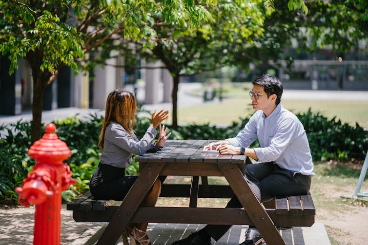 A man and woman sit outside at a picnic table talking in an urban setting, with a red fire hydrant in the foreground. The man takes notes.