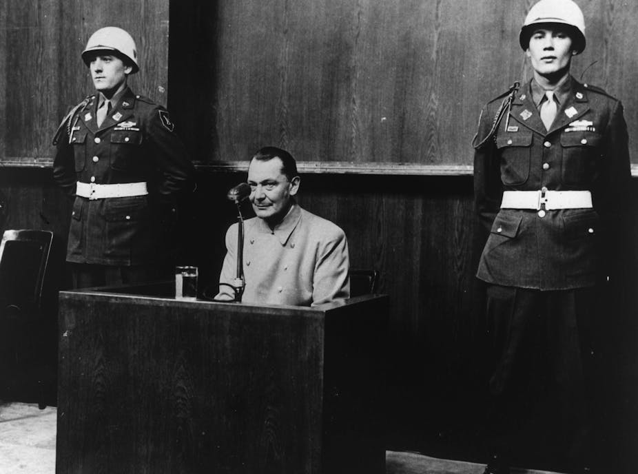 A black and white historical photo showing Herman Goering sitting on the stand in court, with two uniformed military officers beside him, at the Nuremberg tribunals.