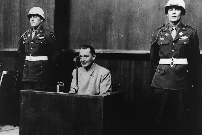 A black and white historical photo showing Herman Goering sitting on the stand in court, with two uniformed military officers beside him, at the Nuremberg tribunals.