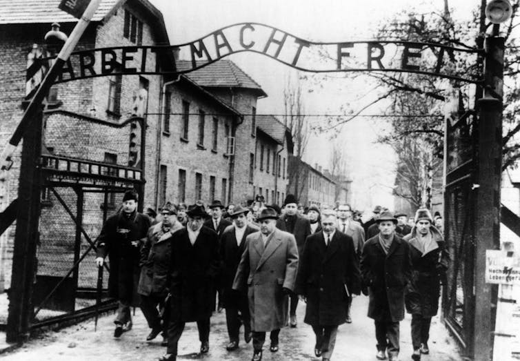Black and white historical photo of a group of men in winter coats and hats walking under the Auschwitz entry sign reading Arbei Macht Frei.