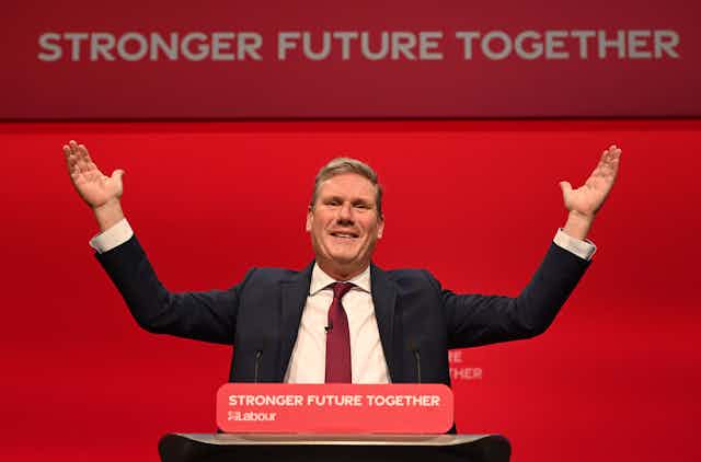 Labour leader Keir Starmer gestures with his hands in the air during his keynote speech at Labour conference. The red background has white text reading Stronger Future Together.