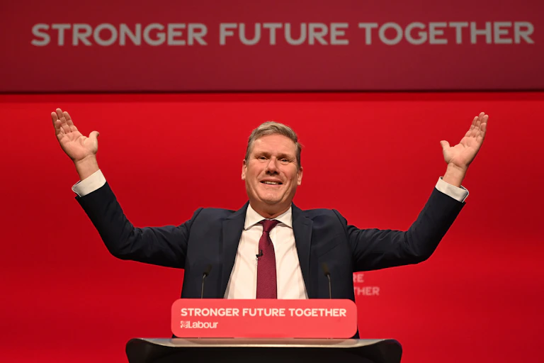 Labour leader Keir Starmer gestures with his hands in the air during his keynote speech at Labour conference. The red background has white text reading Stronger Future Together.